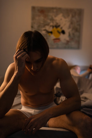 upset and shirtless man sitting on bed near blurred girlfriend, cheating conceptの写真素材