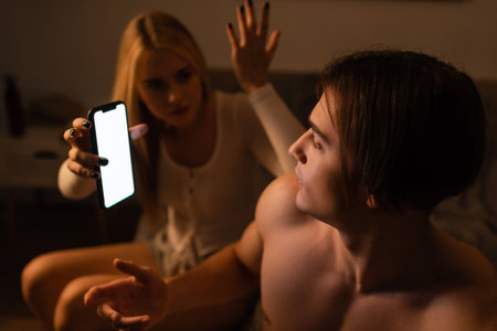 blonde woman holding smartphone while arguing with unfaithful boyfriend at nightの写真素材