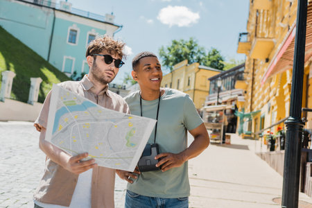 joyful african american tourist with vintage camera looking away near tour guide with city map on...