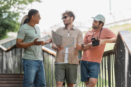 smiling african american man gesturing while talking to friend holding travel map on stairs of urban streetの写真素材