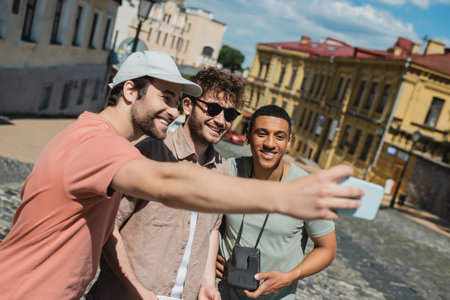 happy tourist in sun cap taking selfie with multiethnic men during excursion on Andrews descent in Kyivの写真素材