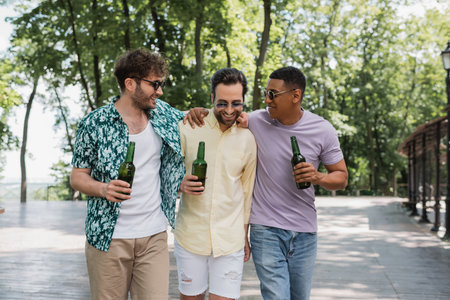 carefree and stylish interracial friends embracing and holding beer during summer walk in city parkの写真素材