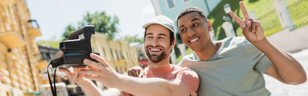 happy african american man showing victory sign near friend taking selfie on vintage camera on...