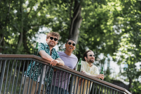 Carefree interracial friends in sunglasses spending time near beer on railing in summer parkの写真素材