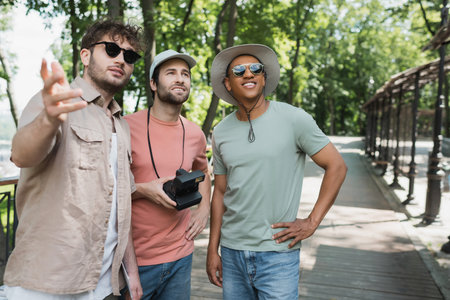 interracial travelers with vintage camera smiling and looking away near tour guide pointing with hand in city parkの写真素材