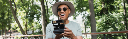 excited african american traveler in sun hat and sunglasses taking picture on vintage camera in...