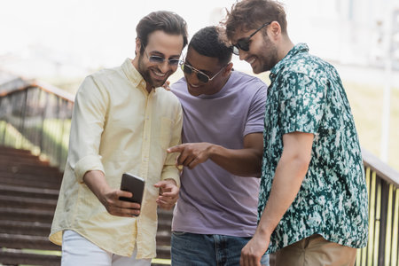 African american man pointing at smartphone near cheerful friends in sunglasses on stairs in parkの写真素材