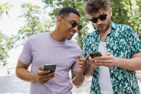 smiling african american man pointing with finger at smartphone in hands of trendy friend in summer parkの写真素材
