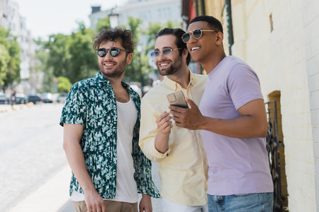 Smiling interracial friends in sunglasses holding cellphone and looking away on street in Kyivの写真素材