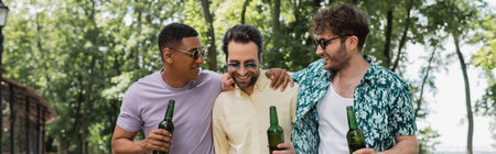 cheerful and trendy multiethnic friends holding beer and embracing while spending time in summer park, bannerの写真素材