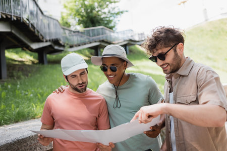 cheerful man in sunglasses pointing at map near interracial friends in sun hats on urban streetの写真素材