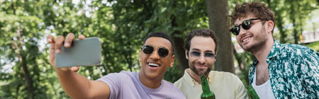 carefree african american man in sunglasses taking selfie with trendy friends holding beer in park, bannerの写真素材
