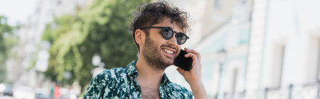 Cheerful young man in sunglasses talking on smartphone while walking on urban street, banner