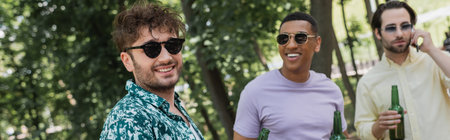 Young man in sunglasses standing near interracial friends with beer in summer park, banner