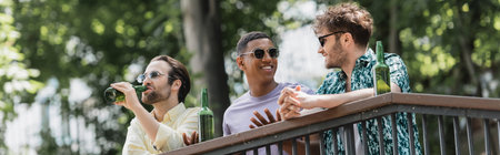 Multiethnic men in sunglasses talking near beer on railing in park, banner