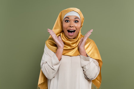Astonished multiracial woman in hijab looking at camera isolated on greenの写真素材