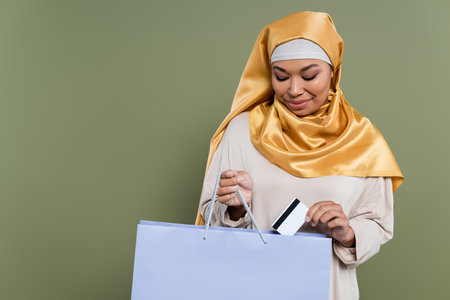 Multiracial woman in hijab putting credit card in shopping bag isolated on greenの写真素材