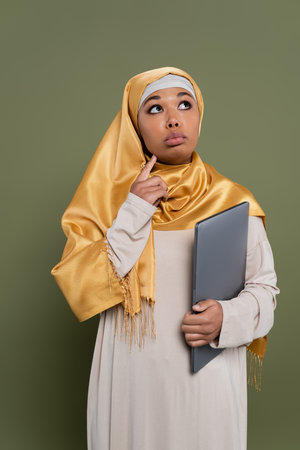 Pensive multiracial woman in hijab holding laptop on green backgroundの写真素材