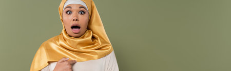 Shocked multiracial woman in yellow hijab pointing with finger at herself isolated on green, bannerの写真素材