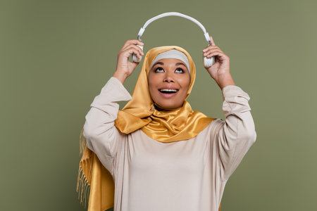 Positive multicultural woman in hijab holding wireless headphones isolated on greenの写真素材