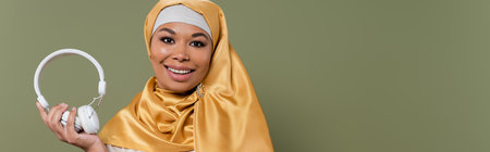 Carefree multiracial woman in hijab holding wireless headphones isolated on green, bannerの写真素材