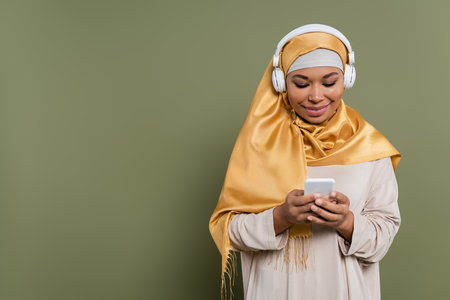 Smiling multiracial woman in hijab and headphones using smartphone on green backgroundの写真素材