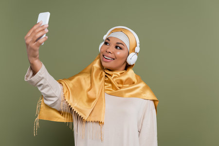Smiling multiracial woman in hijab and headphones taking selfie on smartphone on green backgroundの写真素材