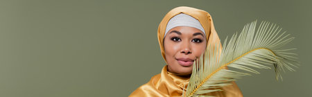 young multiracial woman in yellow silk hijab smiling at camera near leaf of tropical plant isolated on green, bannerの写真素材