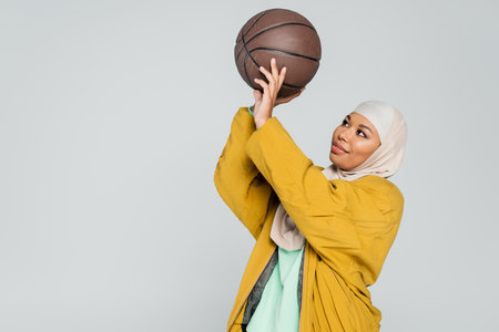 positive multiracial woman in hijab and bomber jacket playing basketball isolated on greyの写真素材