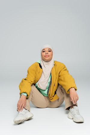 full length of carefree multiracial woman in hijab and jacket with pants sitting on grey backgroundの写真素材