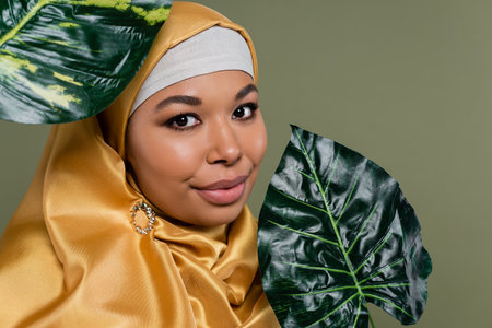 Portrait of multiracial woman in hijab looking at camera near leaves isolated on greenの写真素材