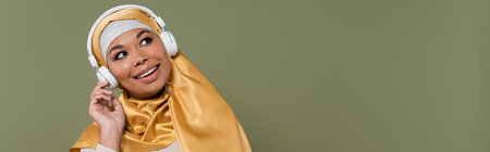Cheerful multiracial woman in hijab listening music in headphones isolated on green, bannerの写真素材