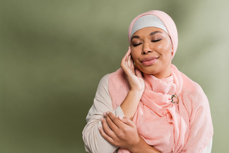 happy multiracial woman in pink hijab posing with closed eyes and touching perfect face on green backgroundの写真素材