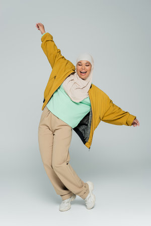 full length of multiracial muslim woman in yellow bomber jacket and beige pants posing with outstretched hands on grey backgroundの写真素材