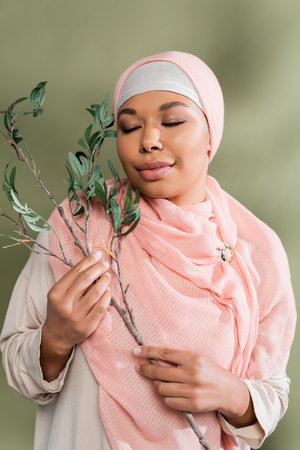 joyful multiracial woman in pink hijab holding olive branch while posing with closed eyes on green...