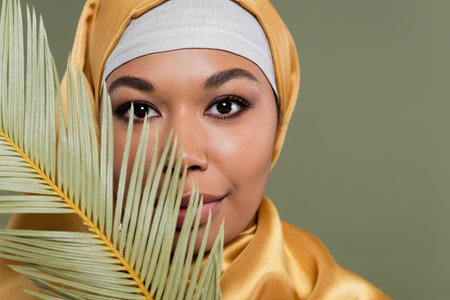 portrait of multiracial muslim woman with makeup looking at camera near tropical leaf isolated on greenの写真素材