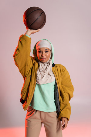stylish multiracial woman in hijab and yellow bomber jacket standing with basketball in raised hand on grey and pink backgroundの写真素材