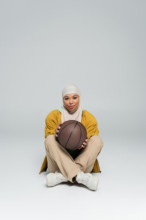 full length of smiling multiracial woman in hijab and pants with sneakers holding basketball while sitting on grey backgroundの写真素材