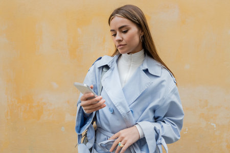 young woman in blue trench coat using smartphone near weathered yellow wall on street in Viennaの写真素材