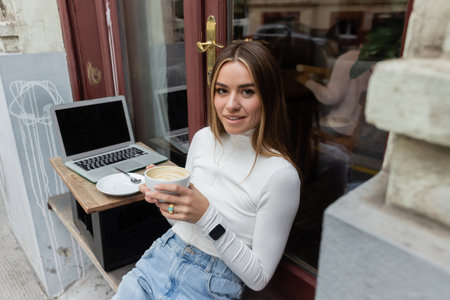 charming woman holding cup of cappuccino while sitting in outdoor cafe next to laptop with blank...