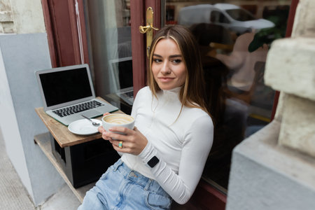 young woman holding cup of cappuccino while sitting in outdoor cafe next to laptop with blank screen...