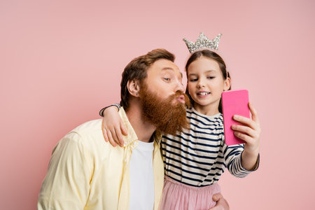 Bearded man pouting lips while taking selfie with daughter isolated on pinkの写真素材