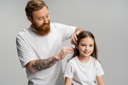 Bearded father adjusting hair of smiling preteen daughter isolated on greyの写真素材