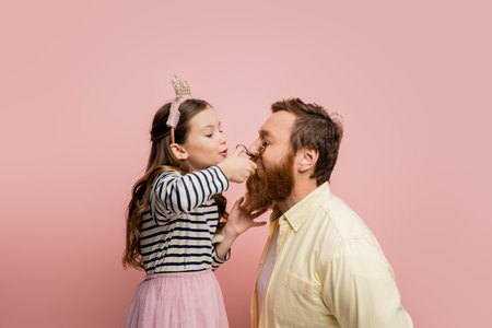Preteen girl with crown headband curling eyelashes of father isolated on pinkの写真素材