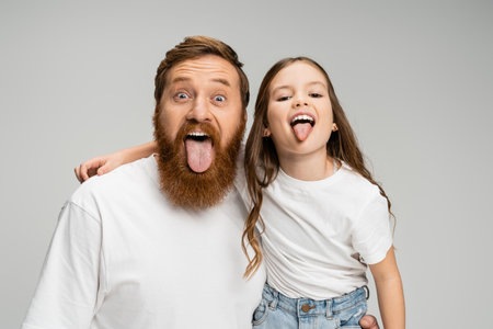 Preteen kid and bearded dad sticking out tongues isolated on greyの写真素材