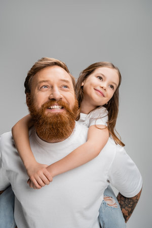 Smiling girl piggybacking on bearded parent and looking away isolated on greyの写真素材