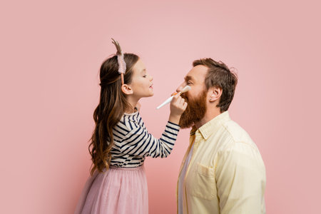 Side view of preteen girl with crown headband holding makeup brush near face of father isolated on pinkの写真素材