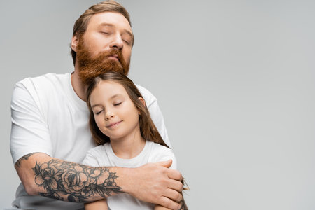 Tattooed man with closed eyes hugging smiling preteen daughter isolated on greyの写真素材