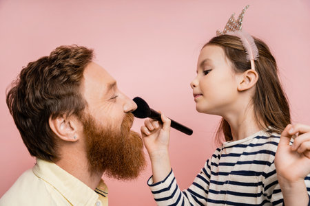 Side view of preteen kid in crown headband applying face powder on bearded father isolated on pinkの写真素材