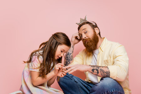 Preteen girl applying nail polish on hand of tattooed dad with crown headband on pink backgroundの写真素材